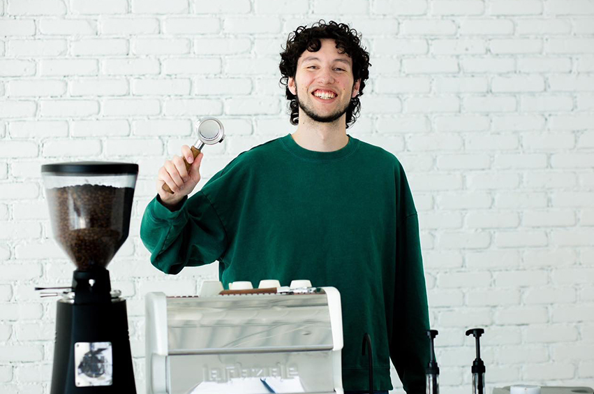 Joseph Arze standing behind a coffee stand in Dallas, Texas, wearing a green sweater